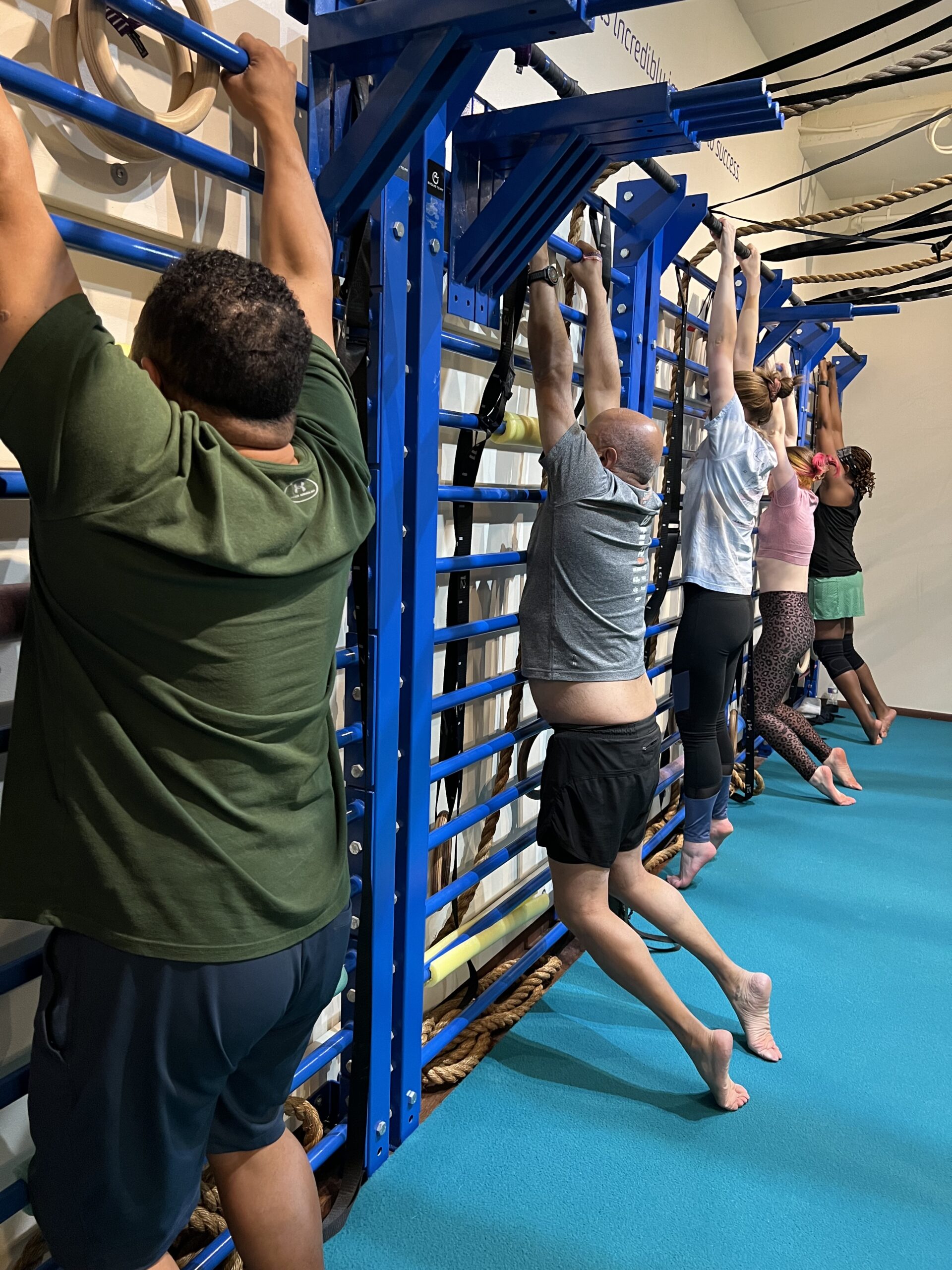 Adults practicing hanging mobility at blue stall bars in the Evolve To Fit studio.